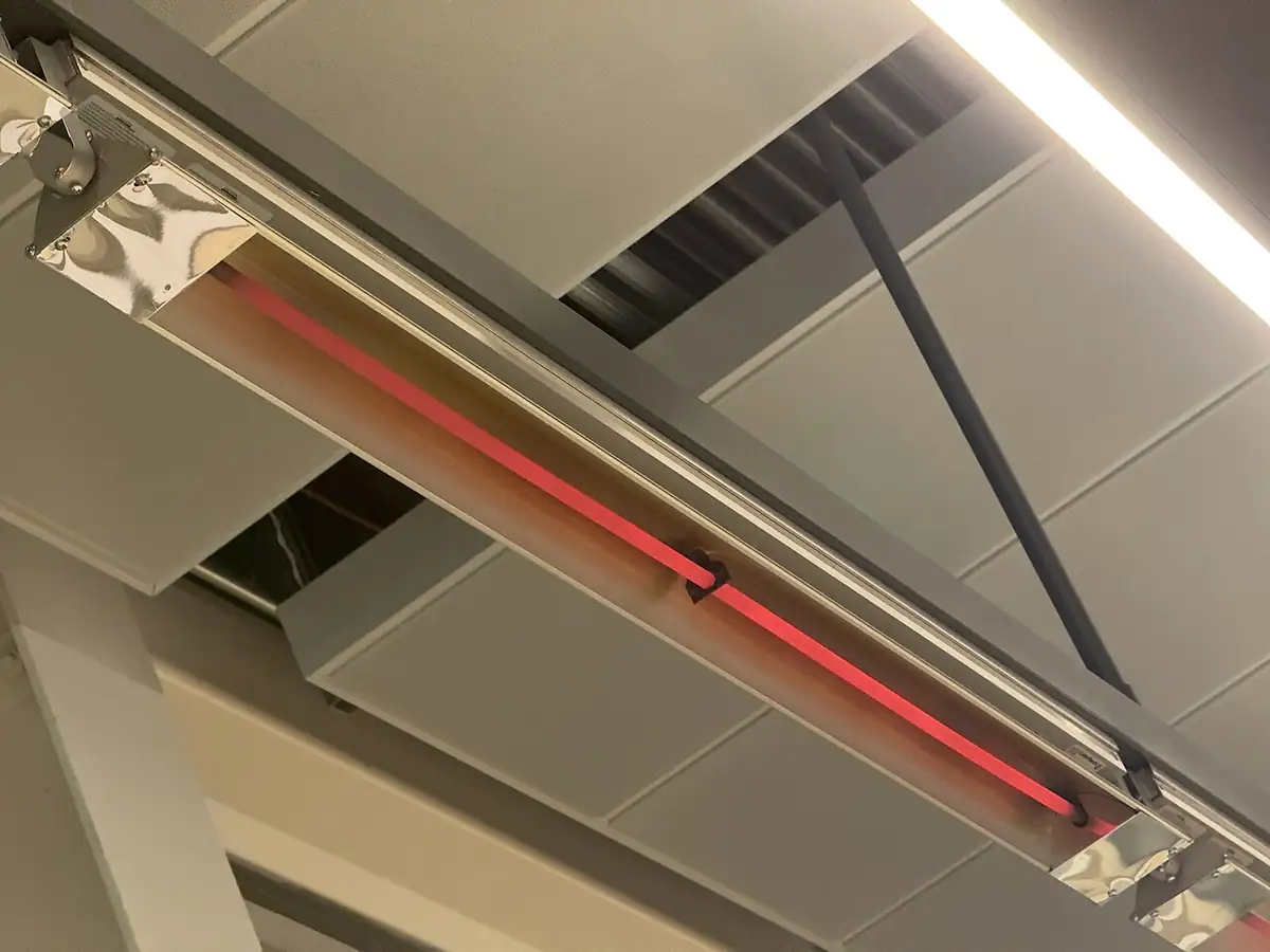 Takmonterad infravärmare med rödglödande element i en blank reflektor, sedd underifrån mot ett innertak
