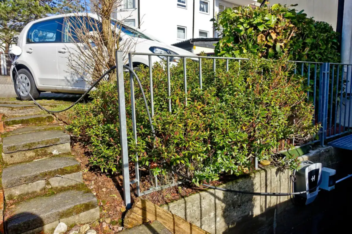 Laddbox på en stödmur vid ett bostadshus, med laddkabeln dragen genom planteringen till en vit elbil på uppfarten
